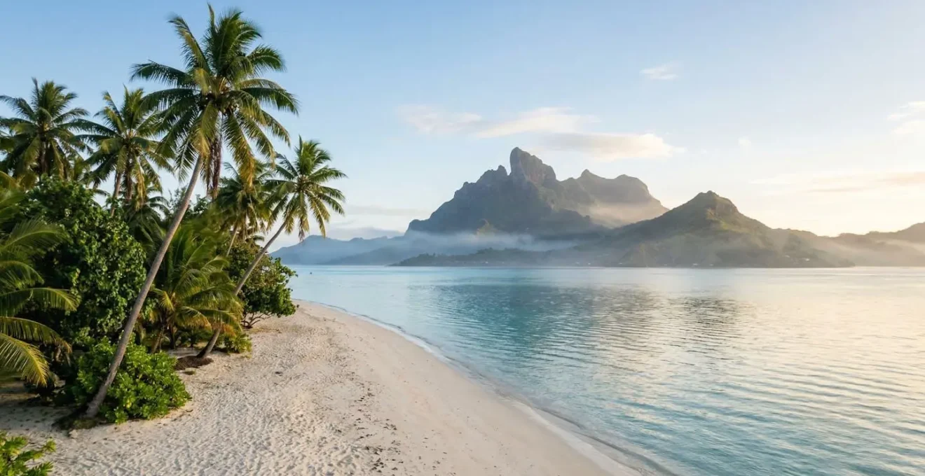 Vue panoramique d'une plage de sable blanc avec lagon turquoise, palmiers et montagne volcanique en arrière-plan sous un ciel azur